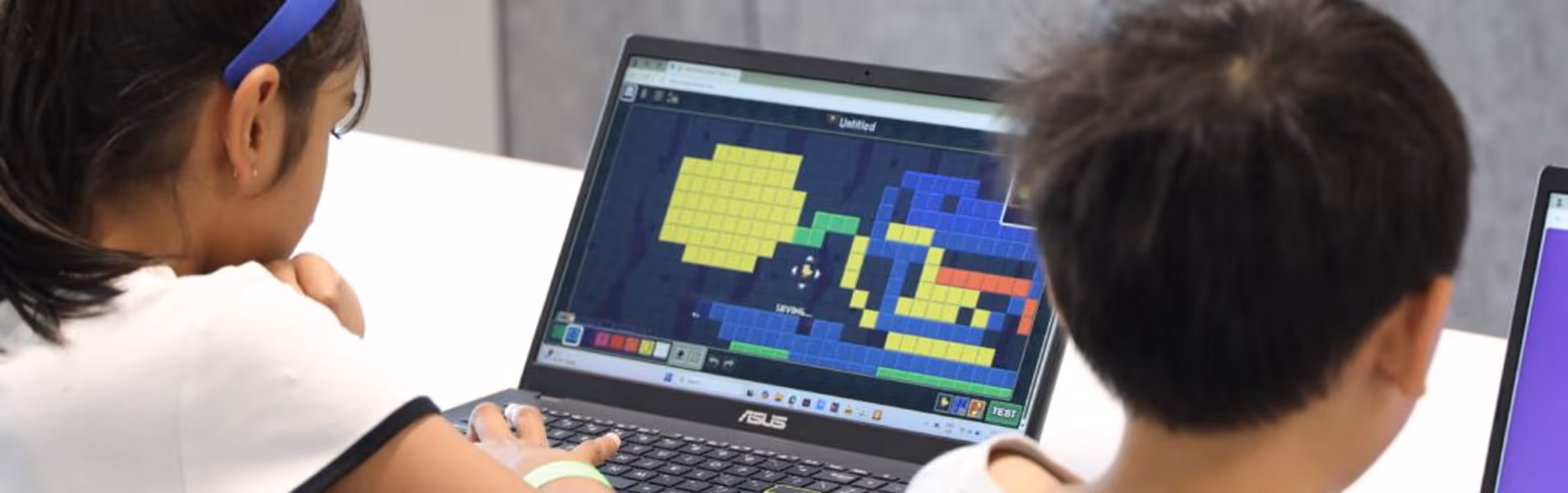 Two children working on laptops; one screen shows pixel art creation using colored blocks.