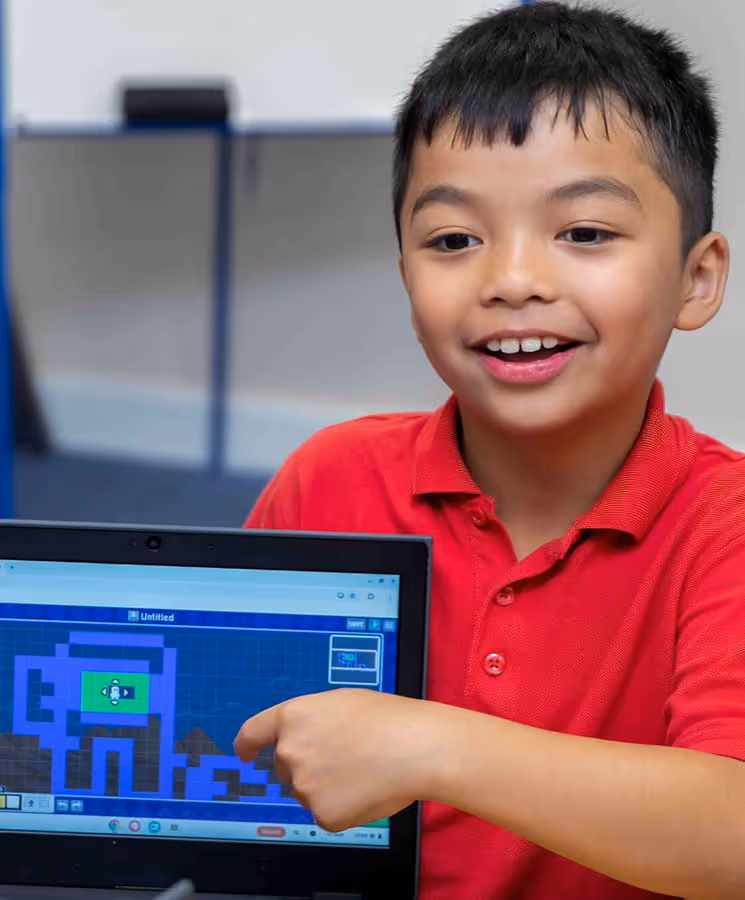 Smiling boy in a red shirt pointing at a laptop screen displaying a pixel art design.
