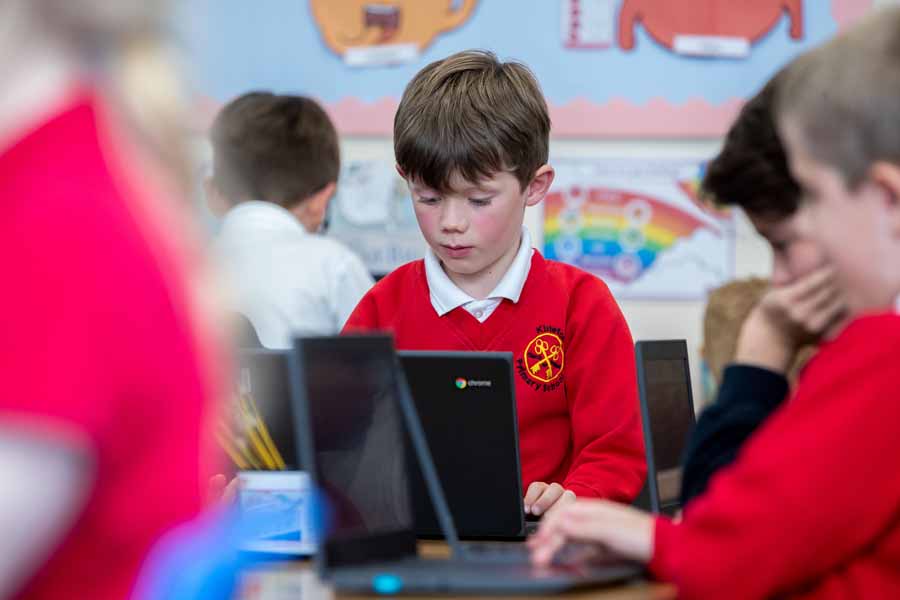Two children seated at desks using computers with a retro-style platform video game displayed on both monitors.
