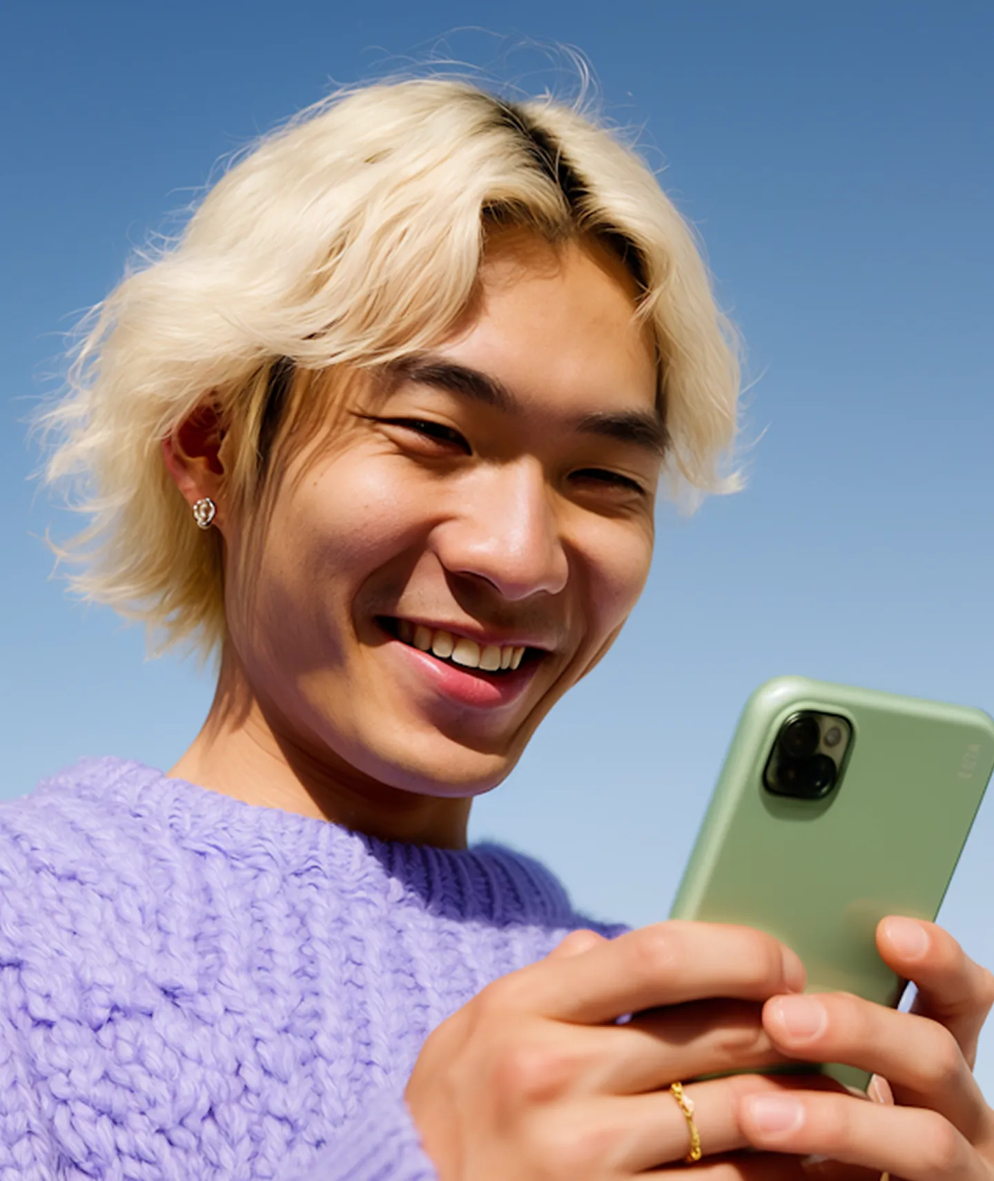 Young person smiling while looking at a green smartphone with clear blue sky background.