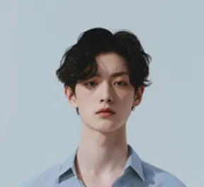Young man with wavy black hair wearing a light blue collared shirt against a pale blue background.