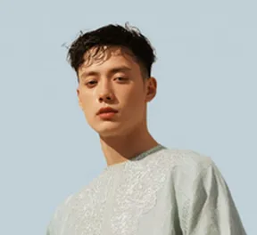 Young man with short dark hair wearing a light blue embroidered top against a plain background.