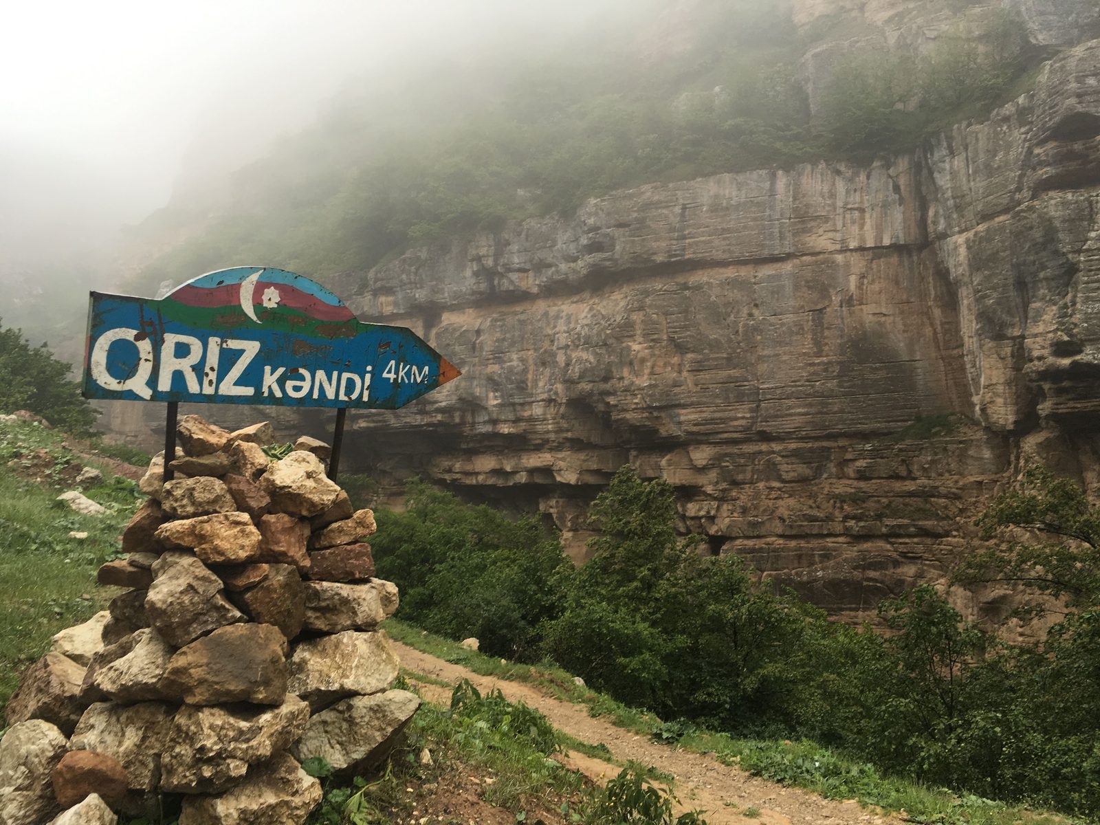 Hand-painted sign to Qrız village on a misted cliff road in the Caucasus — from the founder's archive, 2016.