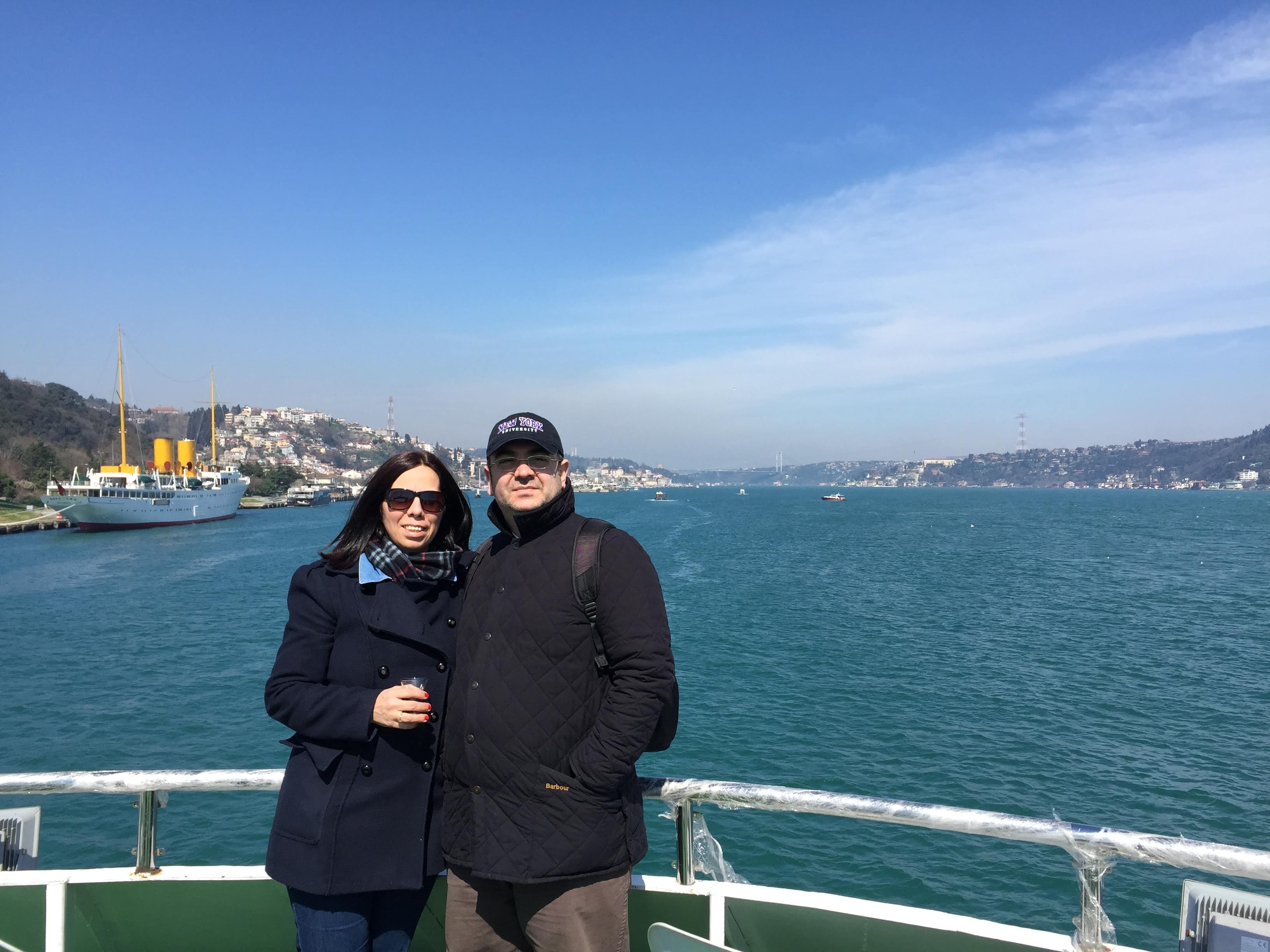 The founder and his wife at the ferry railing on the Bosphorus, the Savarona moored behind them, his New York University cap visible.