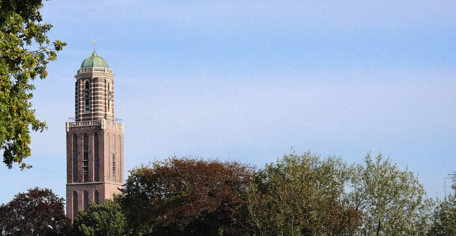 A wide shot of the historic Peperbus tower in Zwolle, Netherlands, rising on the left side above lush green and reddish autumn tree canopies under a clear blue sky.