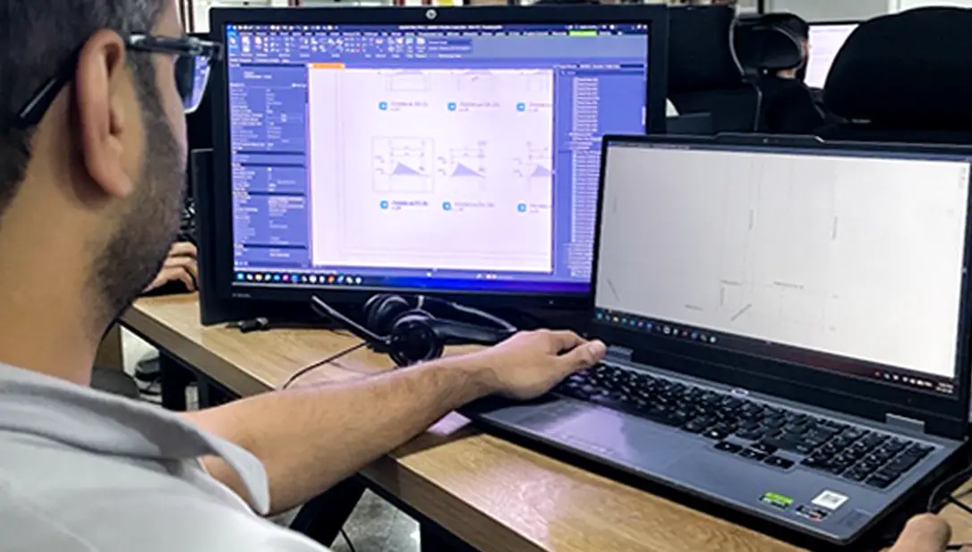 A close-up, over-the-shoulder shot of a technician or engineer wearing glasses, working at a wooden desk with a black laptop open next to a large desktop monitor displaying intricate structural design software blueprints.