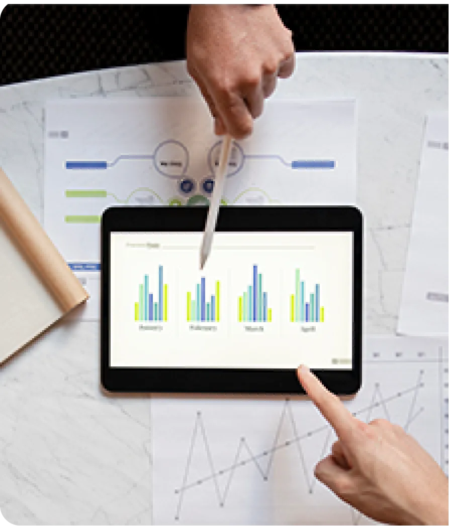 An overhead shot of two people reviewing bar charts on a digital tablet placed over printed data diagrams, with one hand using a stylus and another pointing directly at the screen.