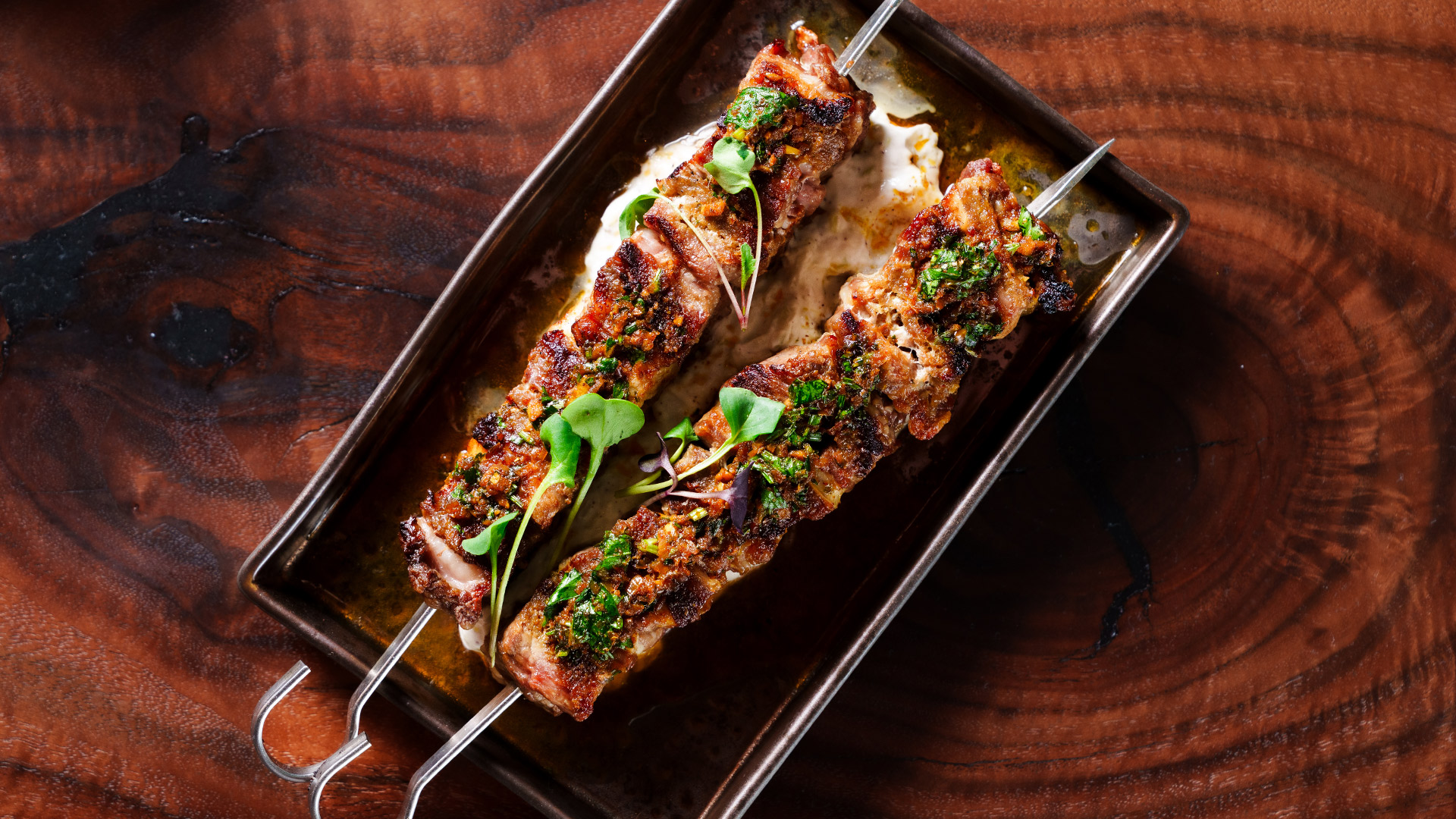 Two grilled meat skewers with herbs on a rectangular dish over a wooden table.