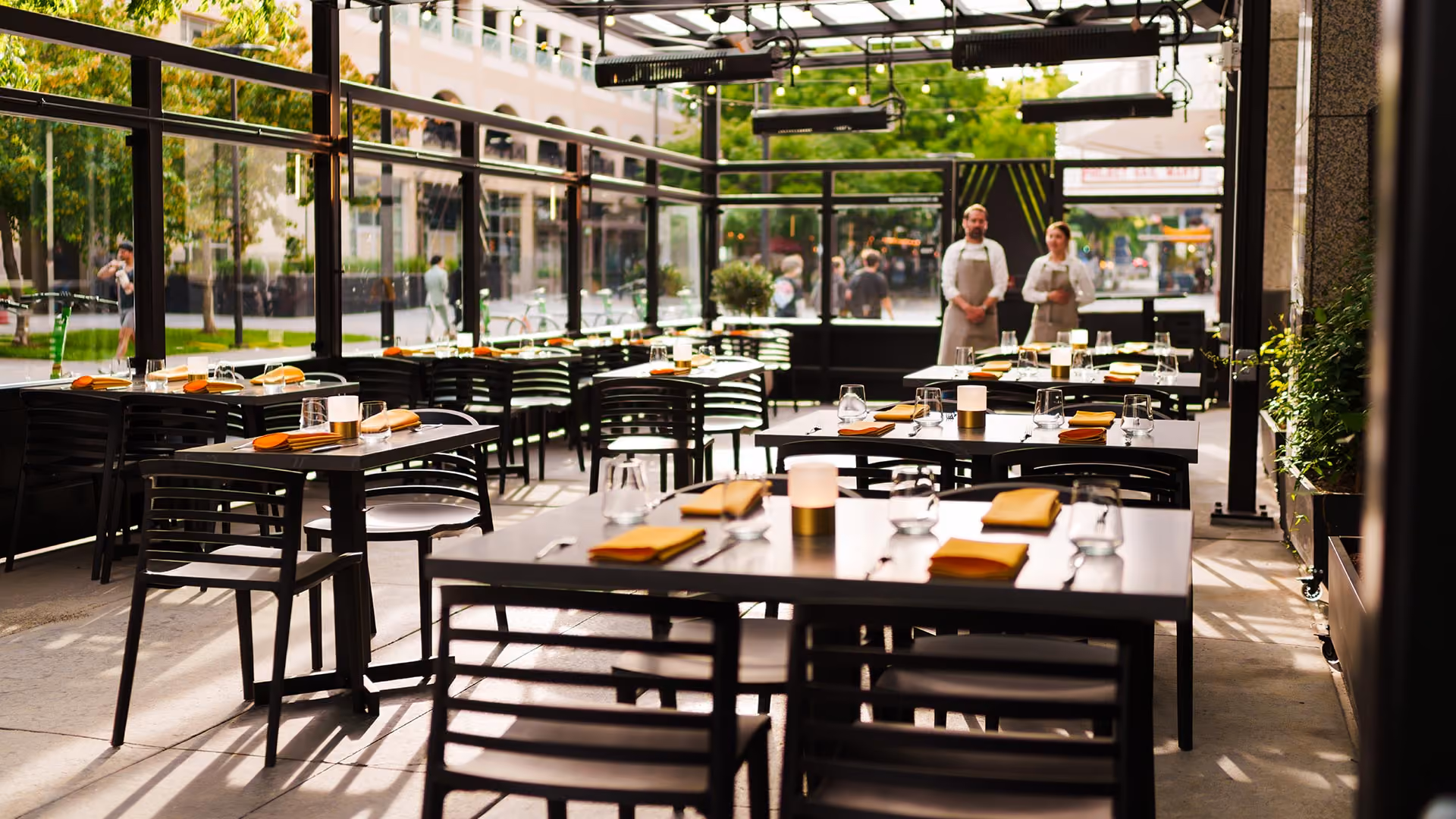 Outdoor restaurant patio with black tables, chairs, orange napkins, glassware, and two waitstaff in aprons.