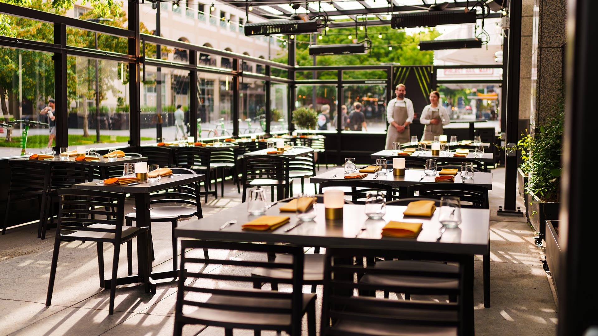 Outdoor restaurant patio with black tables, chairs, orange napkins, glassware, and two waitstaff in aprons.