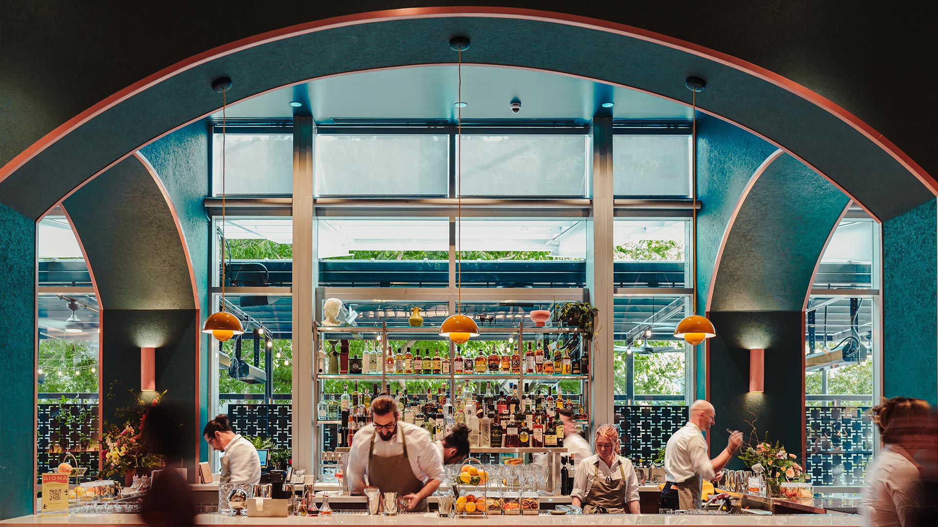 Bartenders preparing drinks behind a well-stocked bar under an arched ceiling with orange pendant lights.