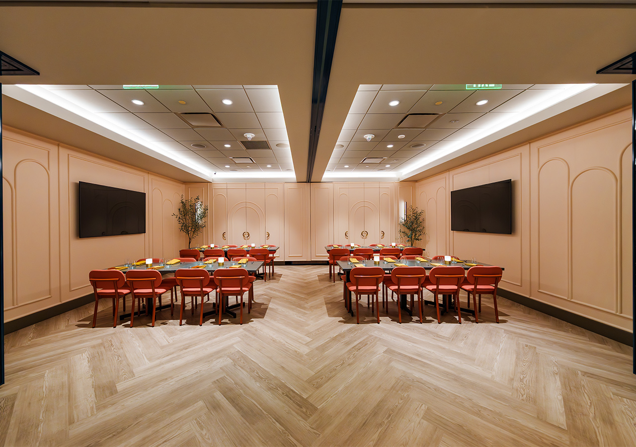 Modern meeting room with two long tables, red chairs, wall-mounted TVs, and wood-patterned floor.