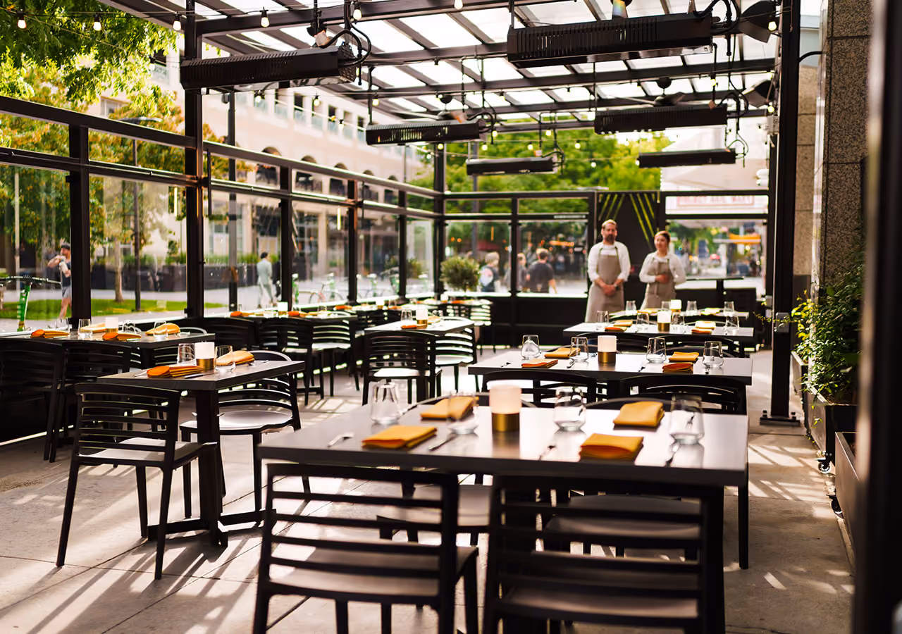 Empty outdoor patio dining area with tables set and two staff standing by the entrance.