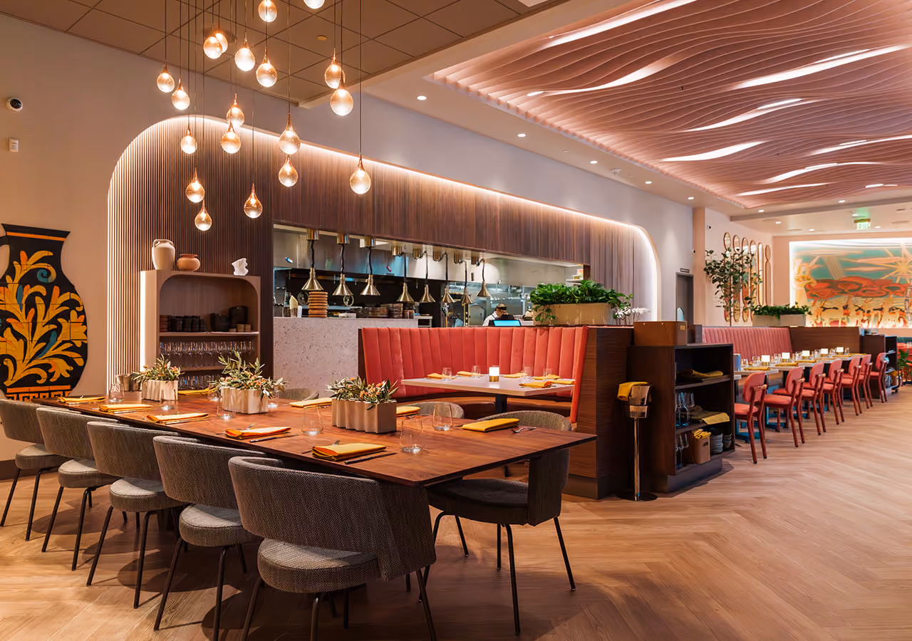Modern restaurant interior with wood tables, red chairs, pendant lights, and open kitchen view.