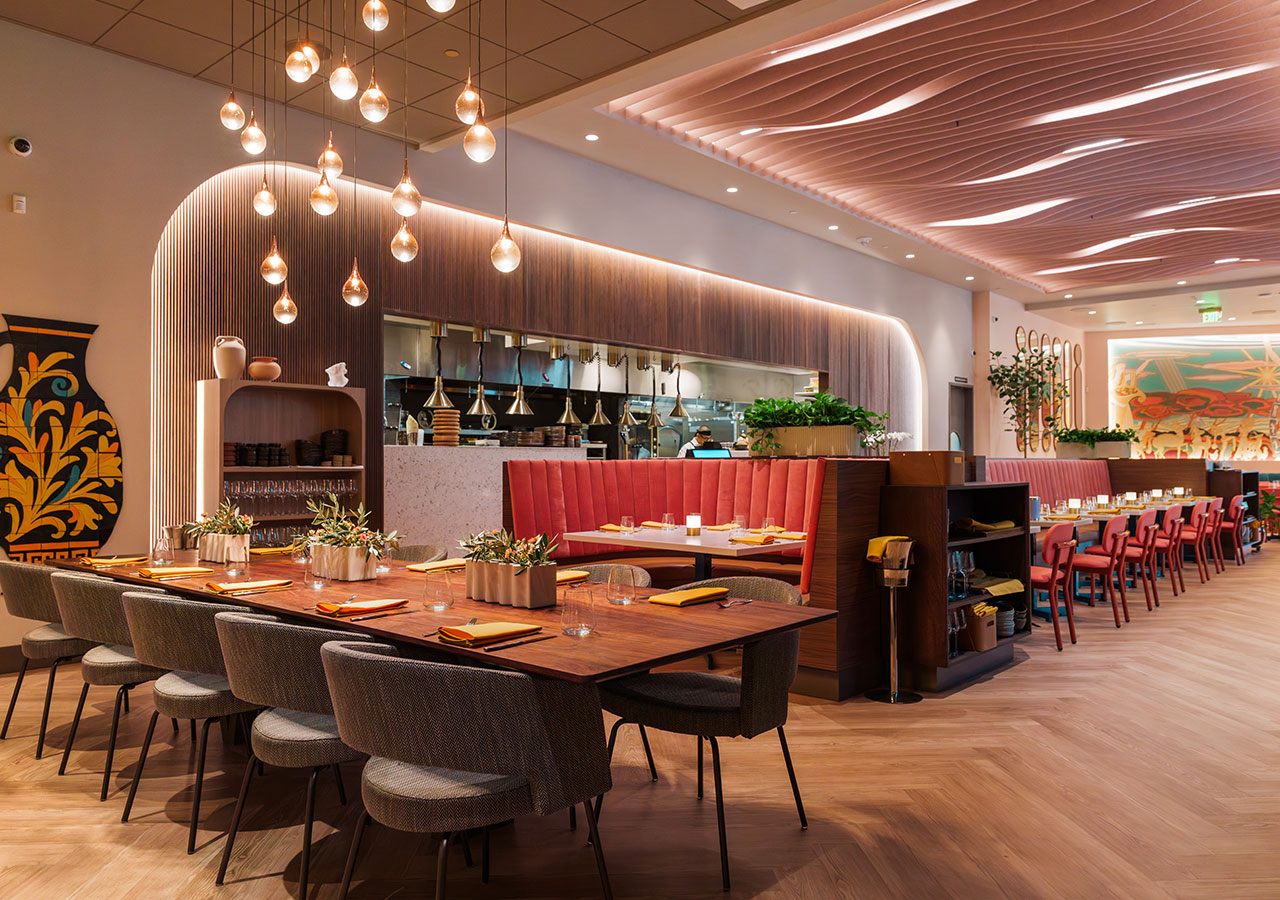 Modern restaurant interior with wood tables, red chairs, pendant lights, and open kitchen view.