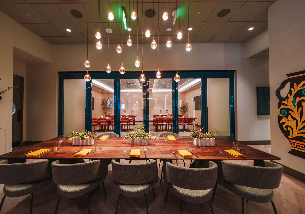 Modern dining room with a wooden table, gray chairs, hanging lights, and a view into another dining area.