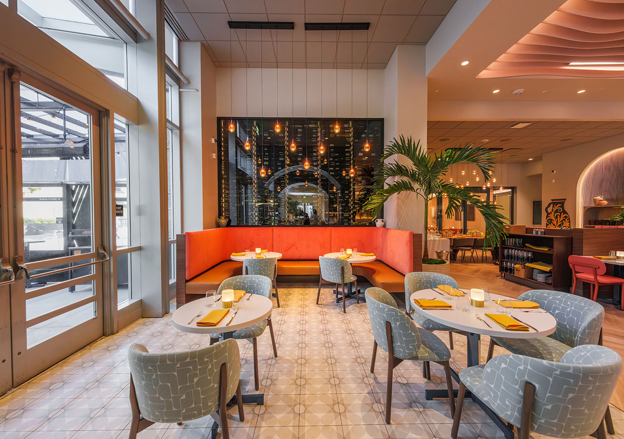 Modern restaurant lounge with orange banquette, patterned chairs, white tables, hanging lights, and large plant.