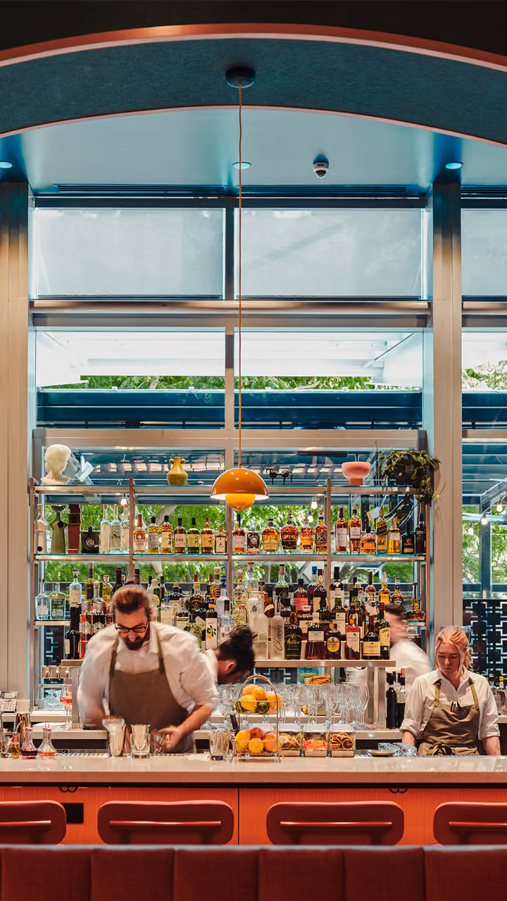 Bartenders preparing drinks behind a bar with shelves of liquor bottles and fruit centerpiece.