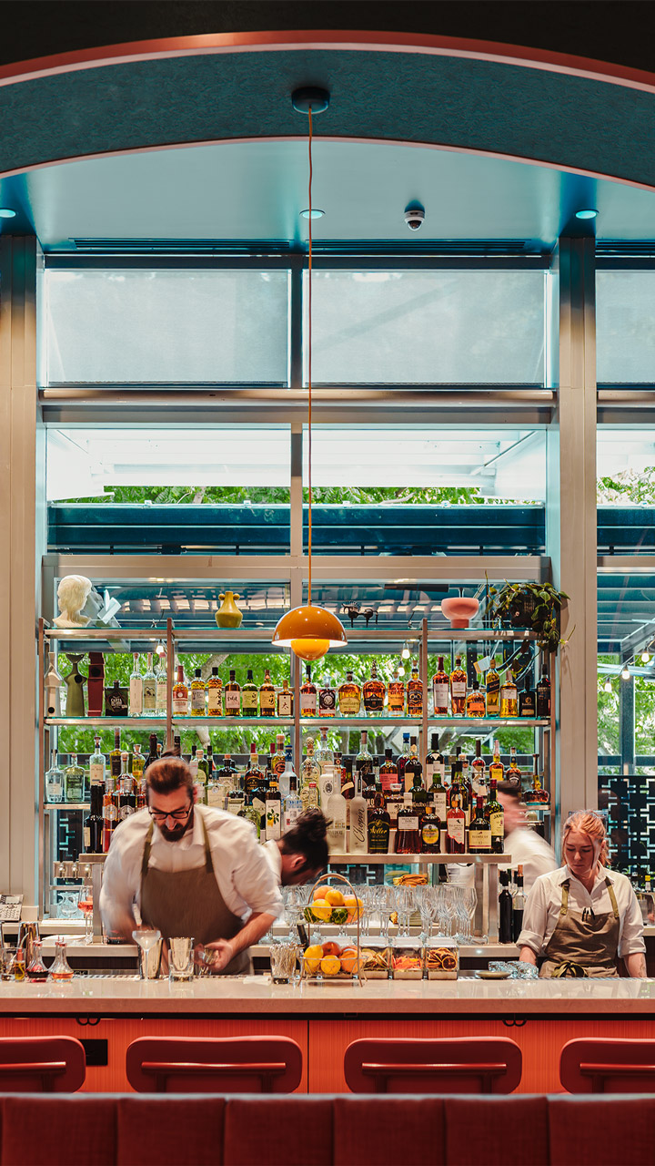 Bartenders preparing drinks behind a bar with shelves of liquor bottles and fruit centerpiece.