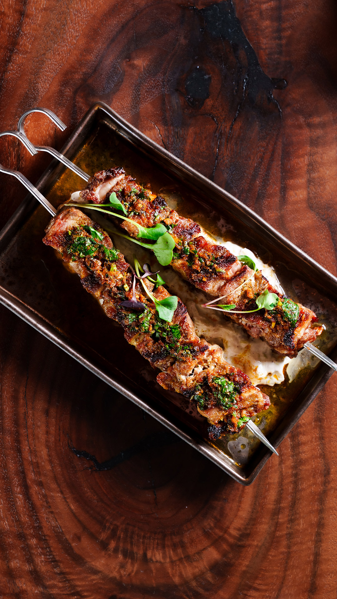 Two grilled meat skewers garnished with herbs, served on a metal tray over sauce.
