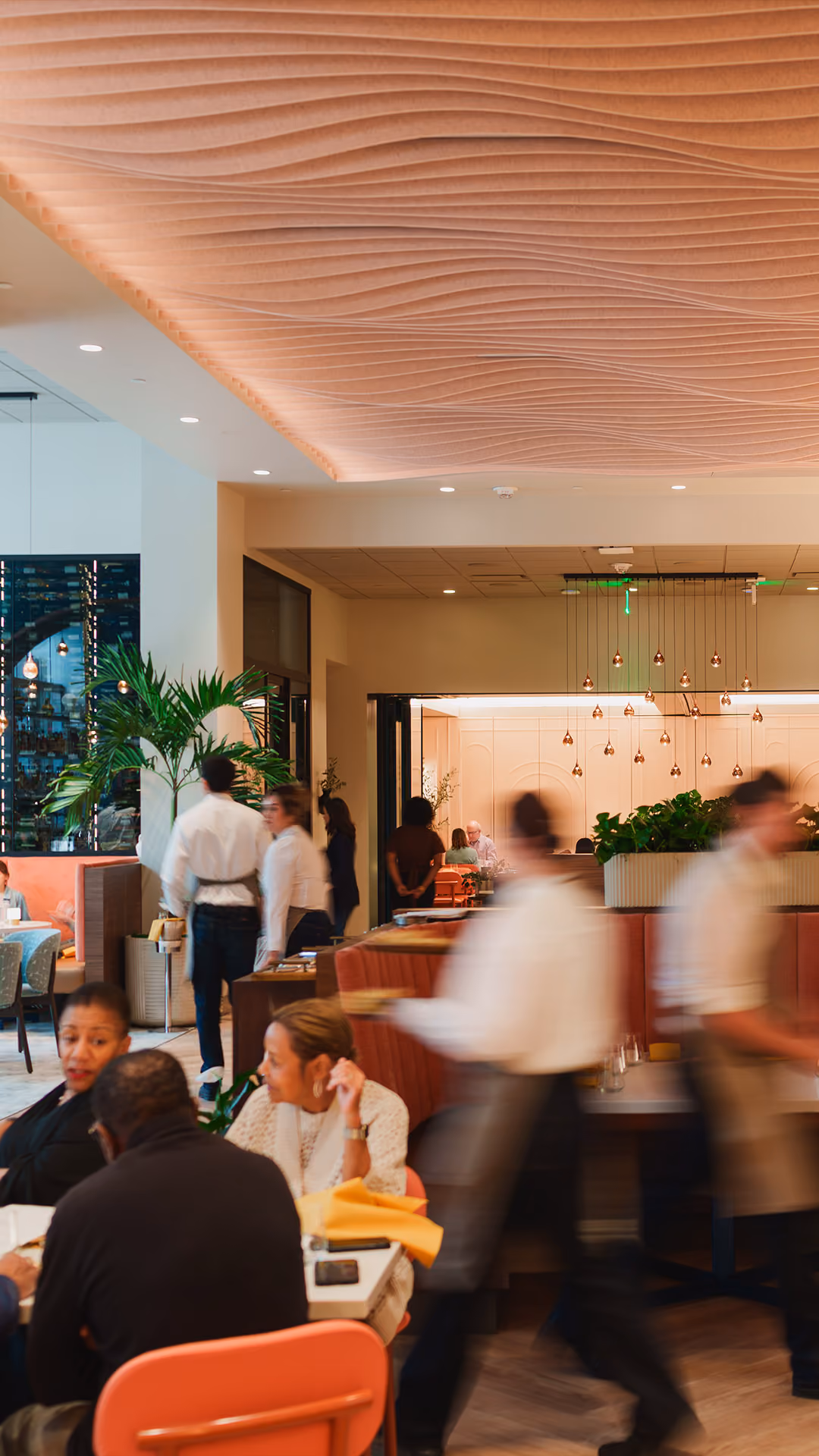 Busy restaurant interior with diners, waitstaff in motion, a plant, and a wavy ceiling design.