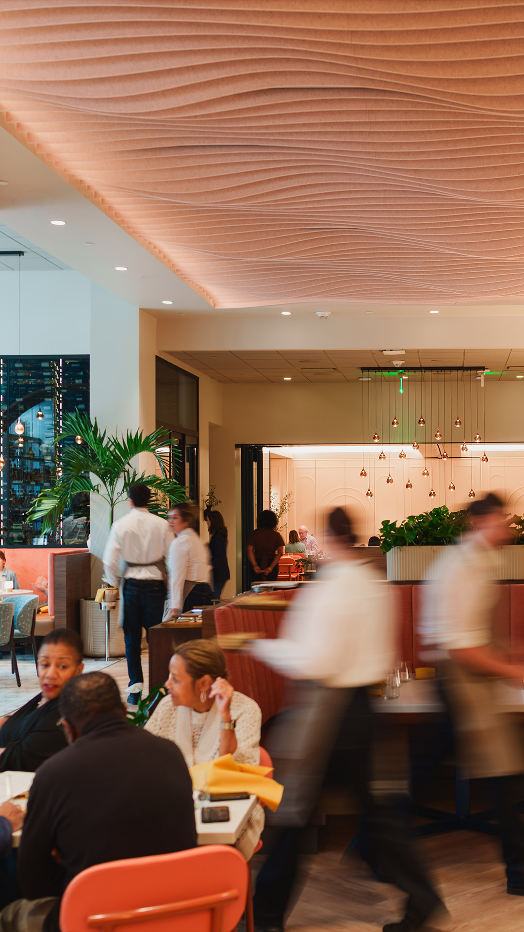 Busy restaurant interior with diners, waitstaff in motion, a plant, and a wavy ceiling design.
