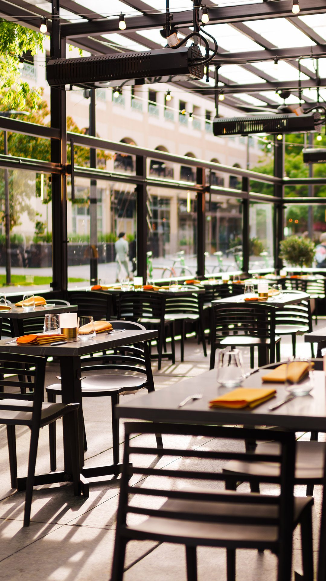 Sunlit outdoor restaurant seating with tables set with orange napkins, glassware, and cutlery.