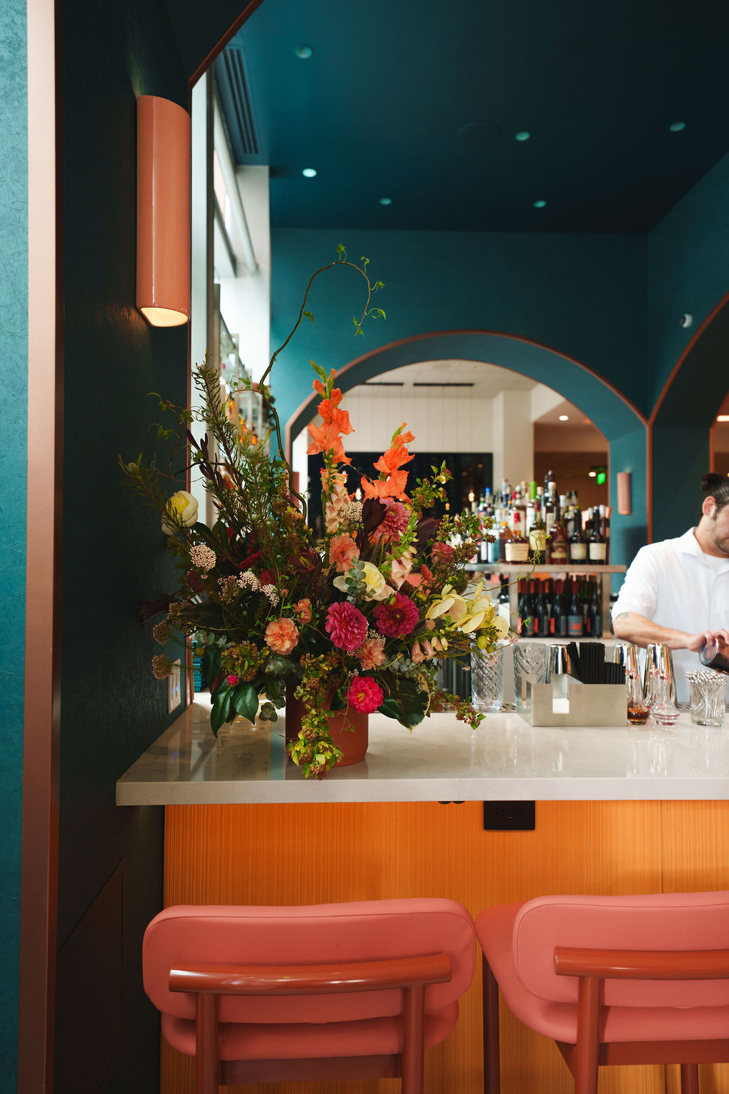 Colorful floral arrangement on bar counter with pink chairs and bartender in background.