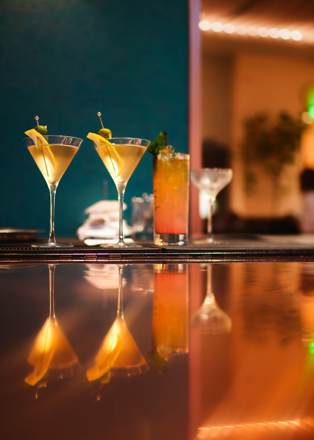 Two martini glasses with olives and lemon peels next to a tall cocktail with mint on a bar counter.