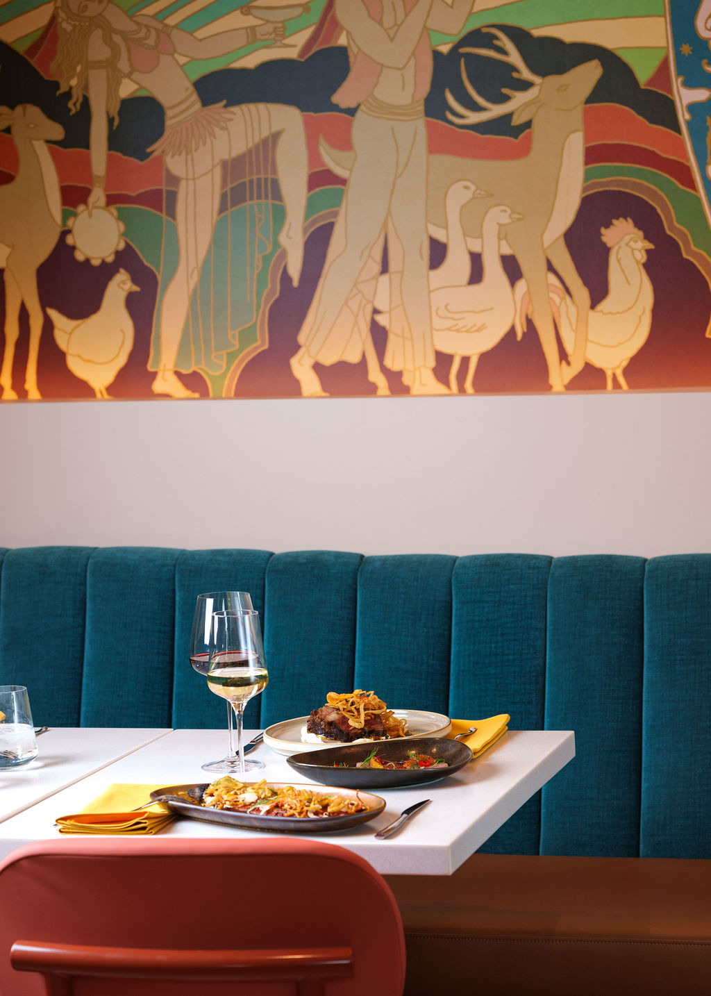 Restaurant table with plates of food and wine glasses against a blue cushioned bench and colorful mural.