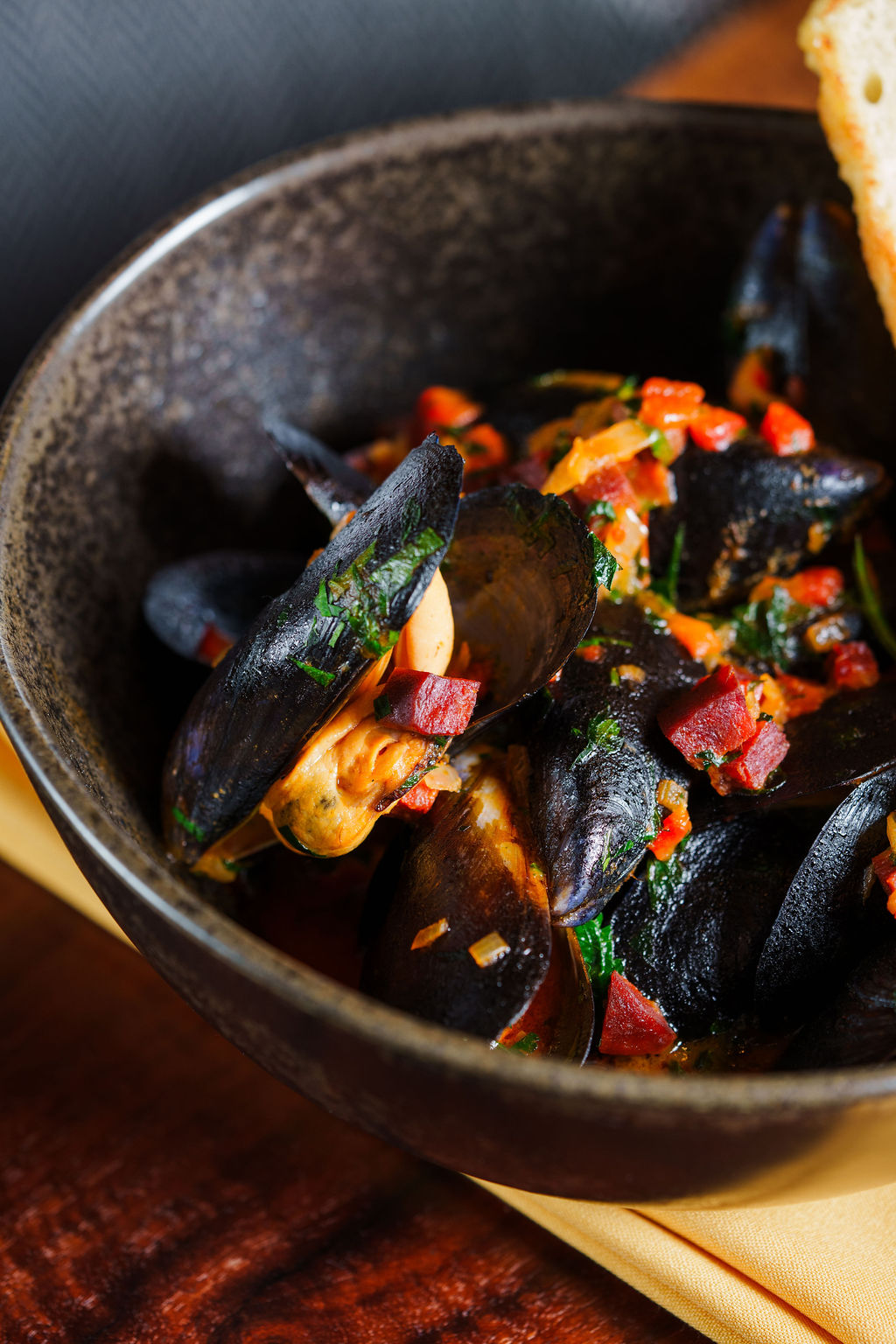 Bowl of cooked mussels garnished with herbs and diced red peppers, with bread slice on the side.