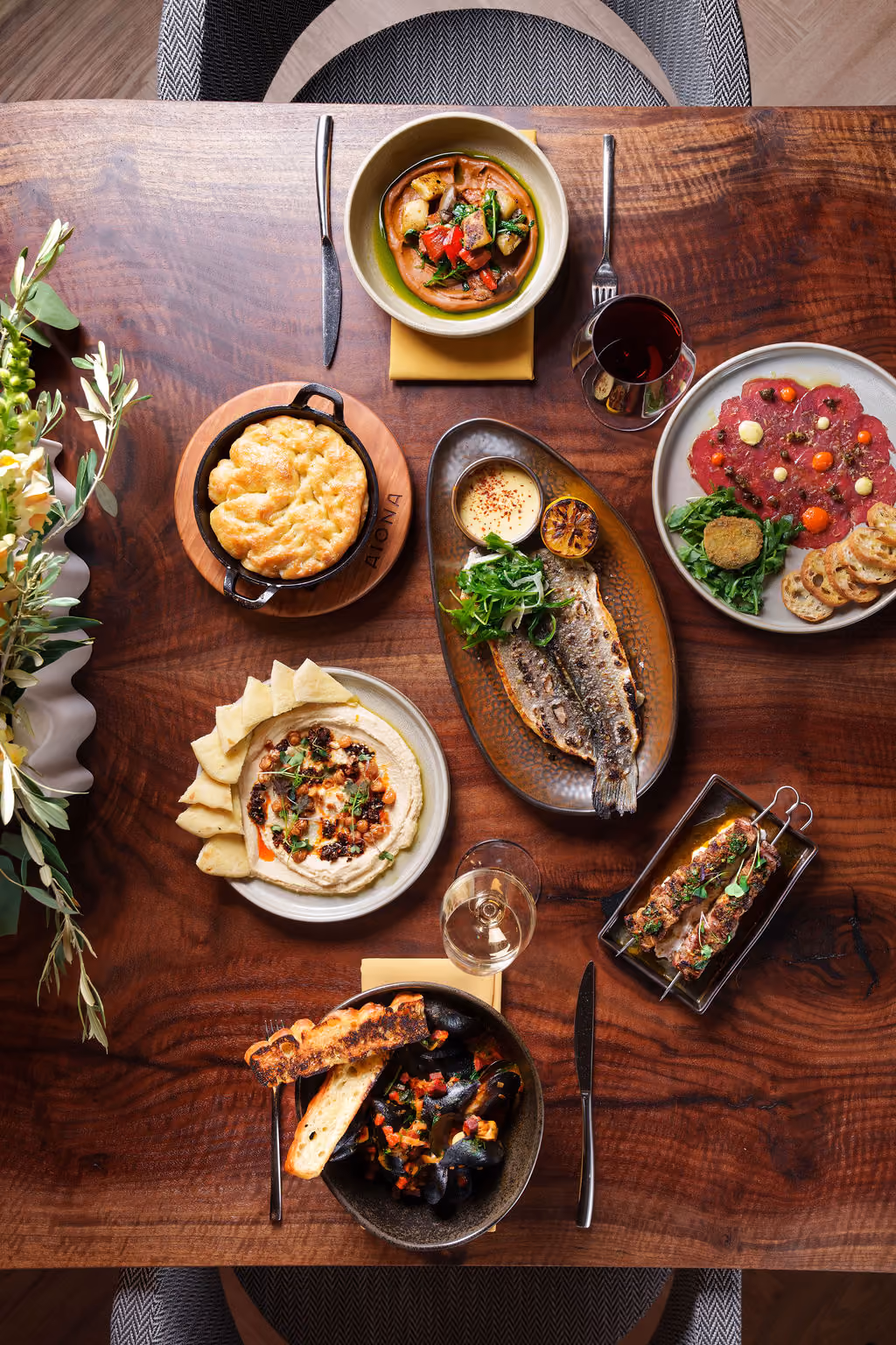 Overhead view of a wooden table with various plated dishes including fish, hummus, bread, and wine glasses.