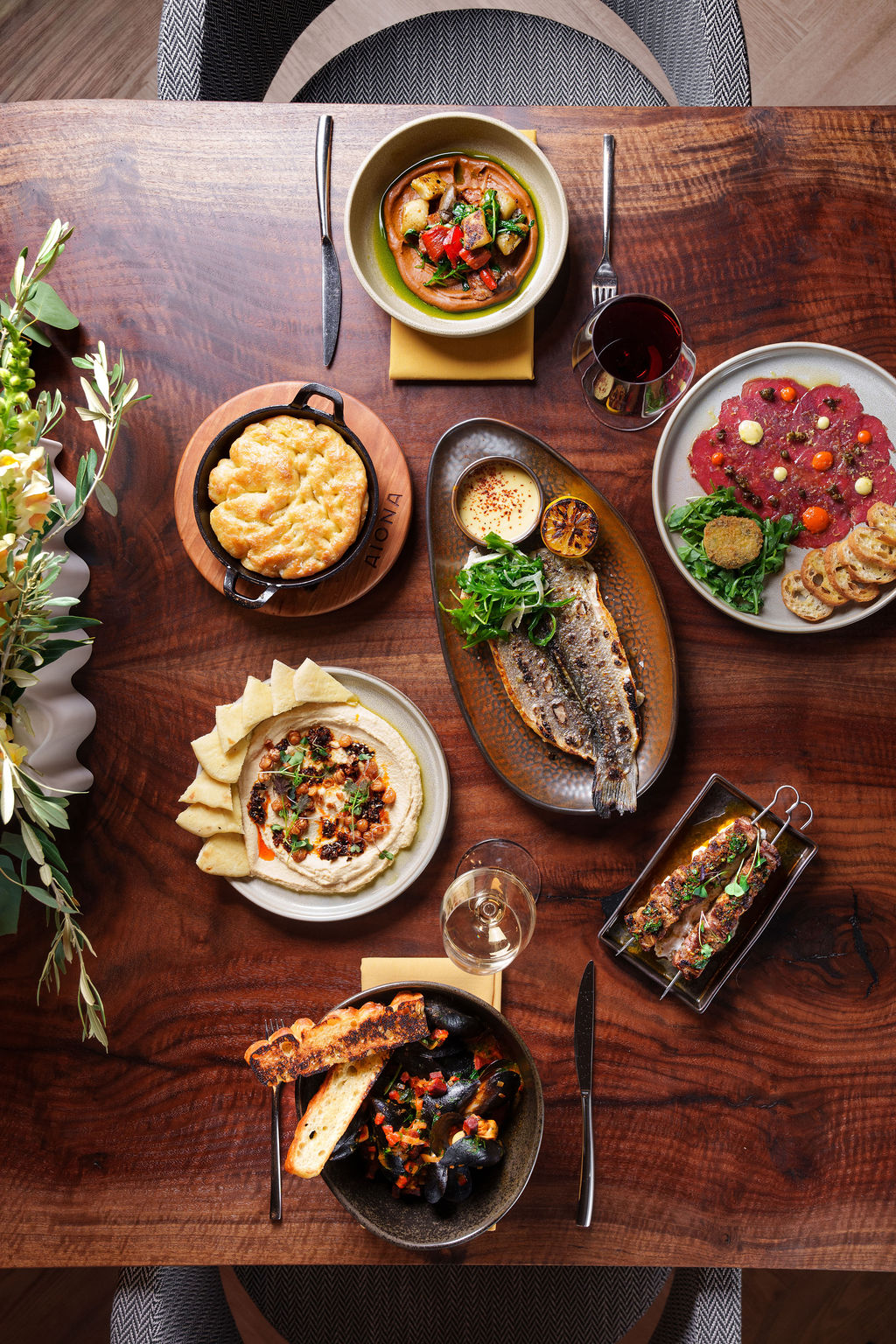 Overhead view of a wooden table with various plated dishes including fish, hummus, bread, and wine glasses.