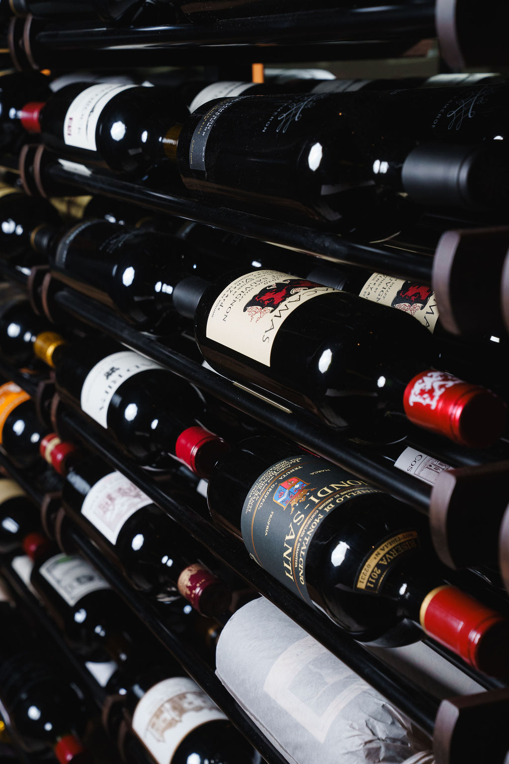 Multiple bottles of red wine stored horizontally on metal racks in a dark wine cellar.