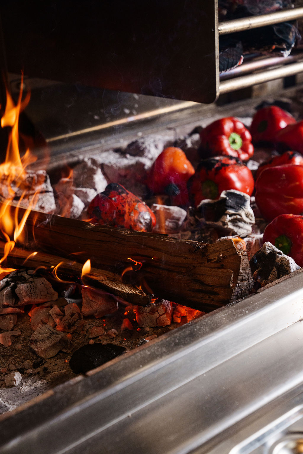 Red bell peppers roasting on hot coals with burning wood and flames in a metal grill.