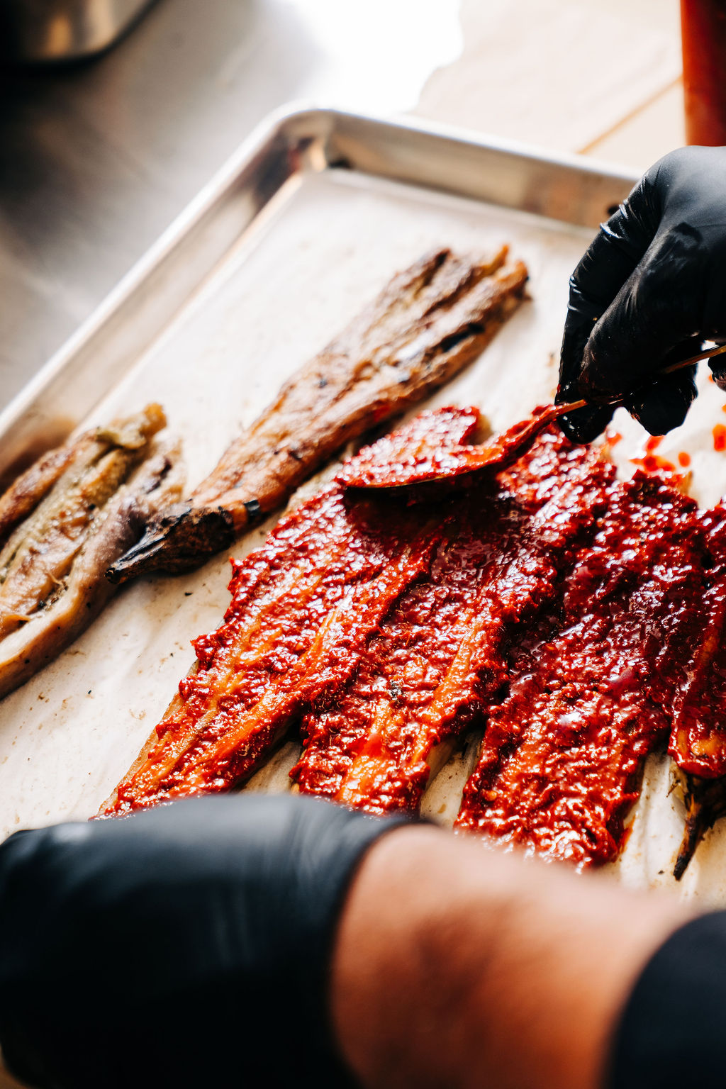 Hands with black gloves spreading red sauce on grilled fish fillets on a metal tray.