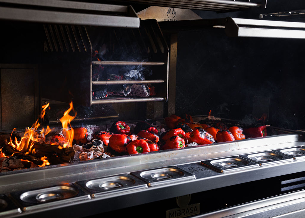 Red bell peppers roasting on an open flame grill with glowing embers and flames.