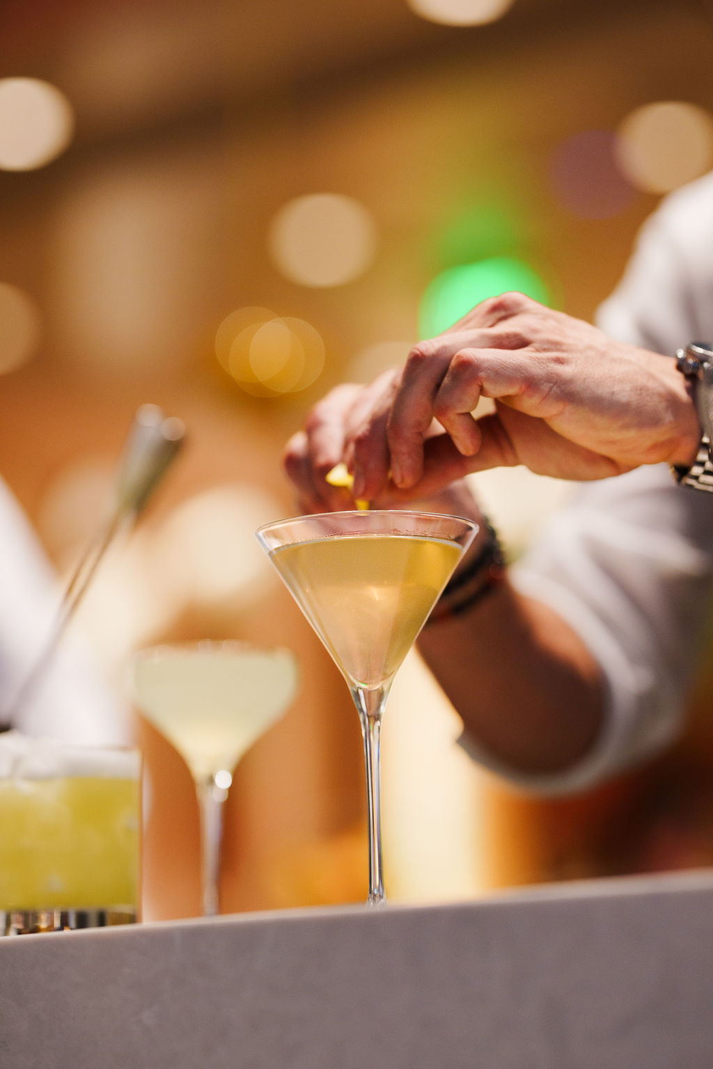 A person garnishing a light yellow cocktail in a martini glass at a bar counter.