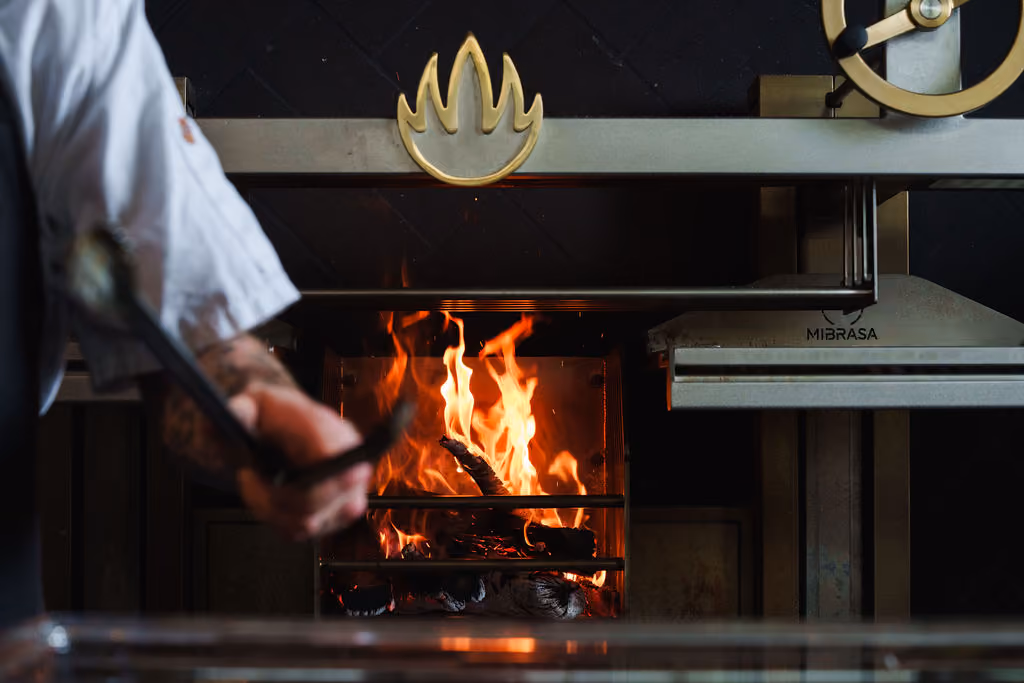 Chef tending to a wood fire inside a stainless steel commercial oven with a flame emblem above it.