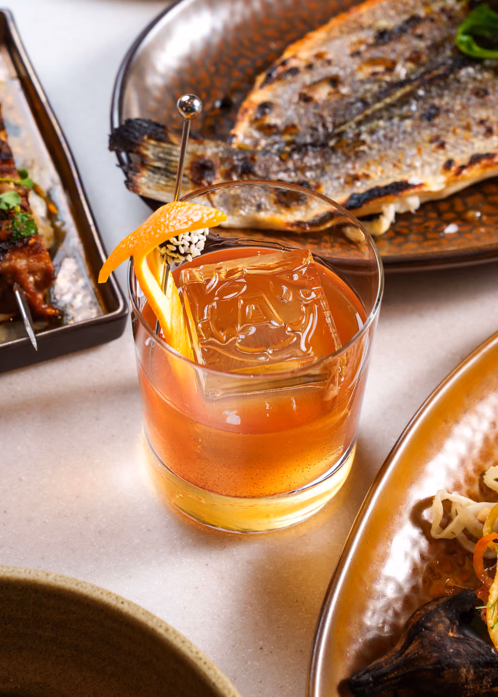 Close-up of a cocktail with an orange peel garnish and large ice cube on a table with grilled fish nearby.