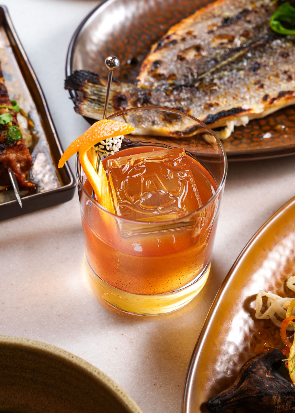 Close-up of a cocktail with an orange peel garnish and large ice cube on a table with grilled fish nearby.