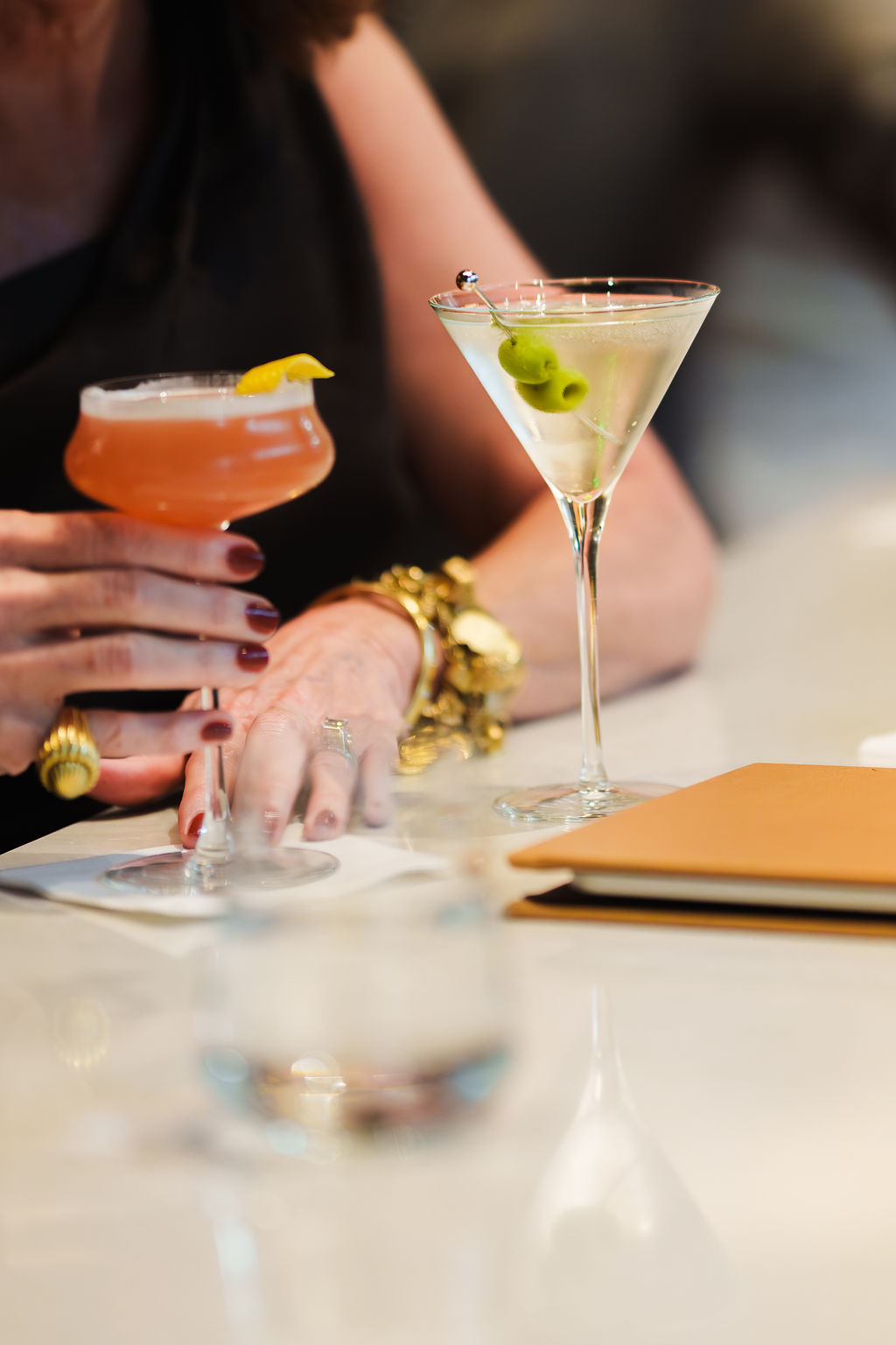 Woman holding a cocktail with a lemon peel next to a martini glass garnished with green olives.