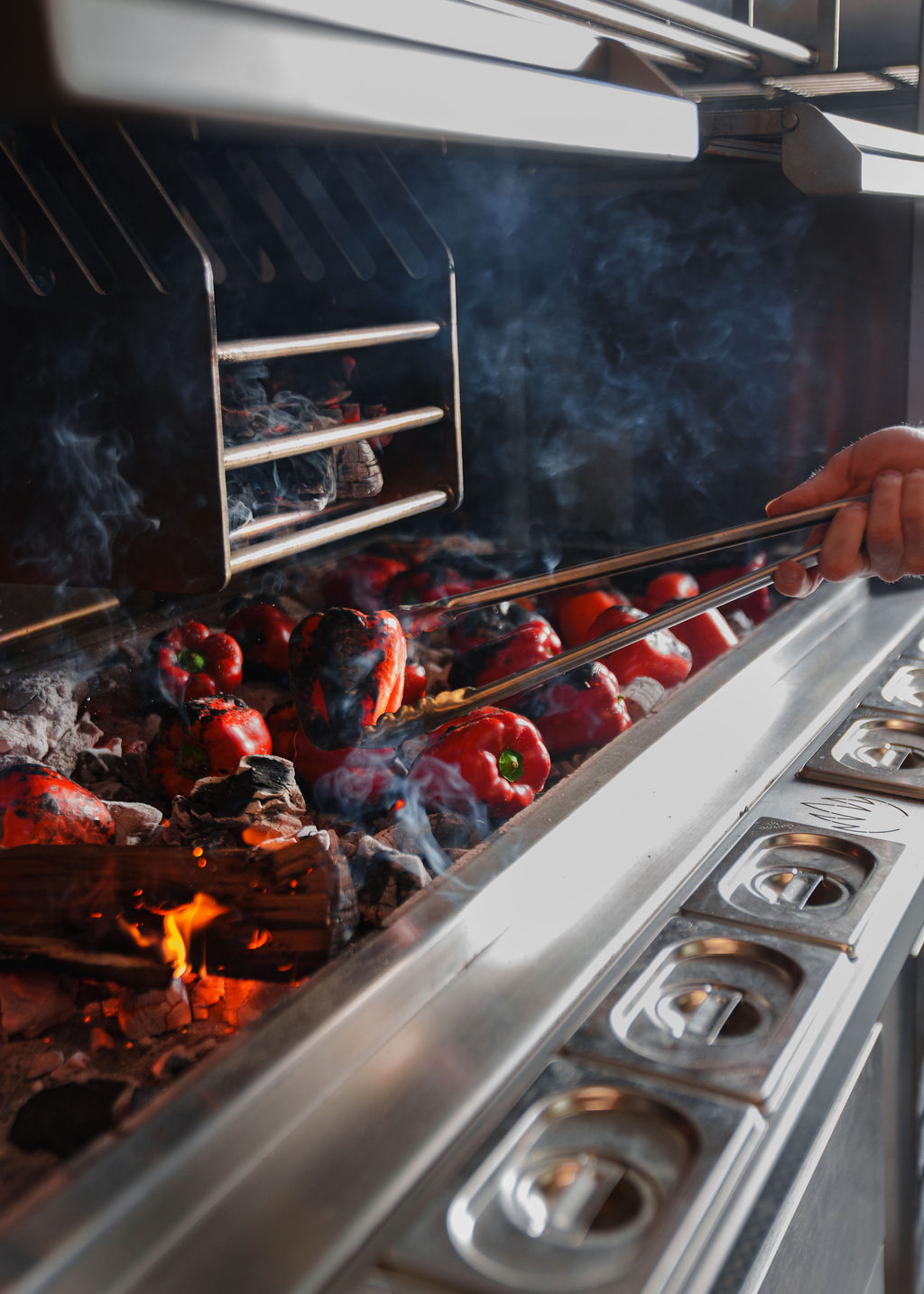 Red bell peppers being grilled over open flames on a charcoal grill with metal tongs.