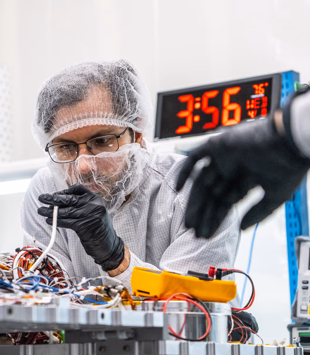 Technician in a lab with hairnet and gloves working on complex electronic wiring and components.