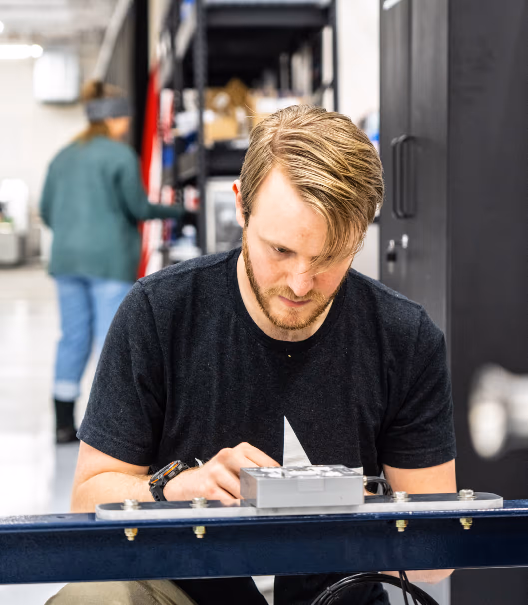 Man with blond hair working on an electronic device on a blue metal surface in a workshop.