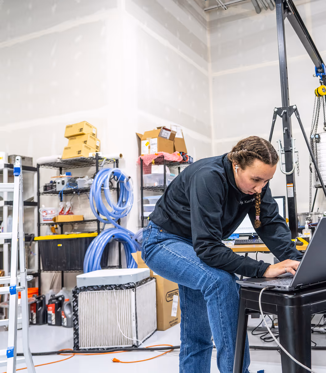 Woman with braids works on laptop in a workshop filled with tools, coils, and equipment.