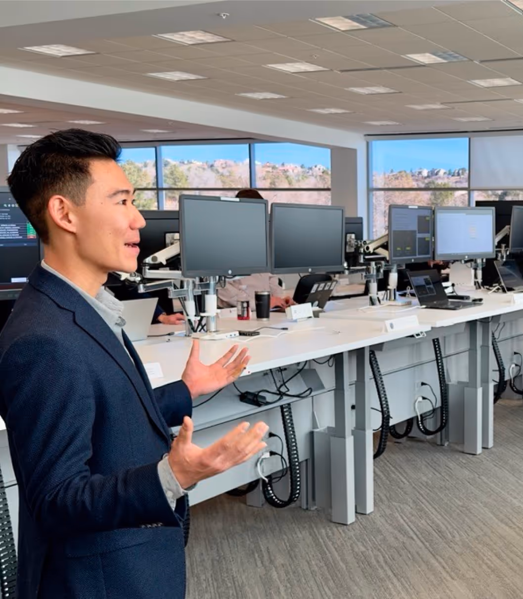 Man in blazer speaking in a modern office with desks, monitors, and people working on laptops.