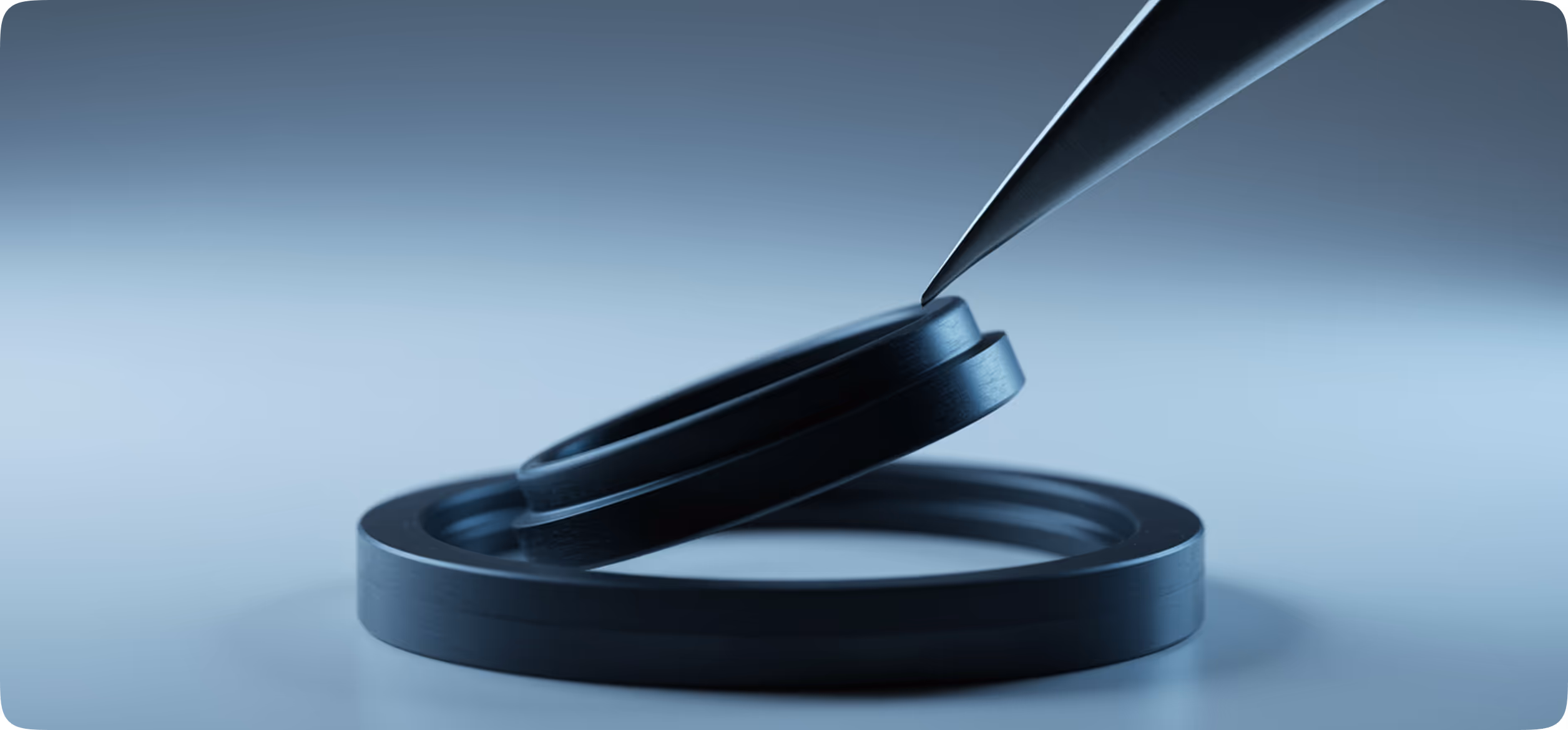 Thin black precision tweezers holding a small black circular ring above a larger black ring on a smooth surface.