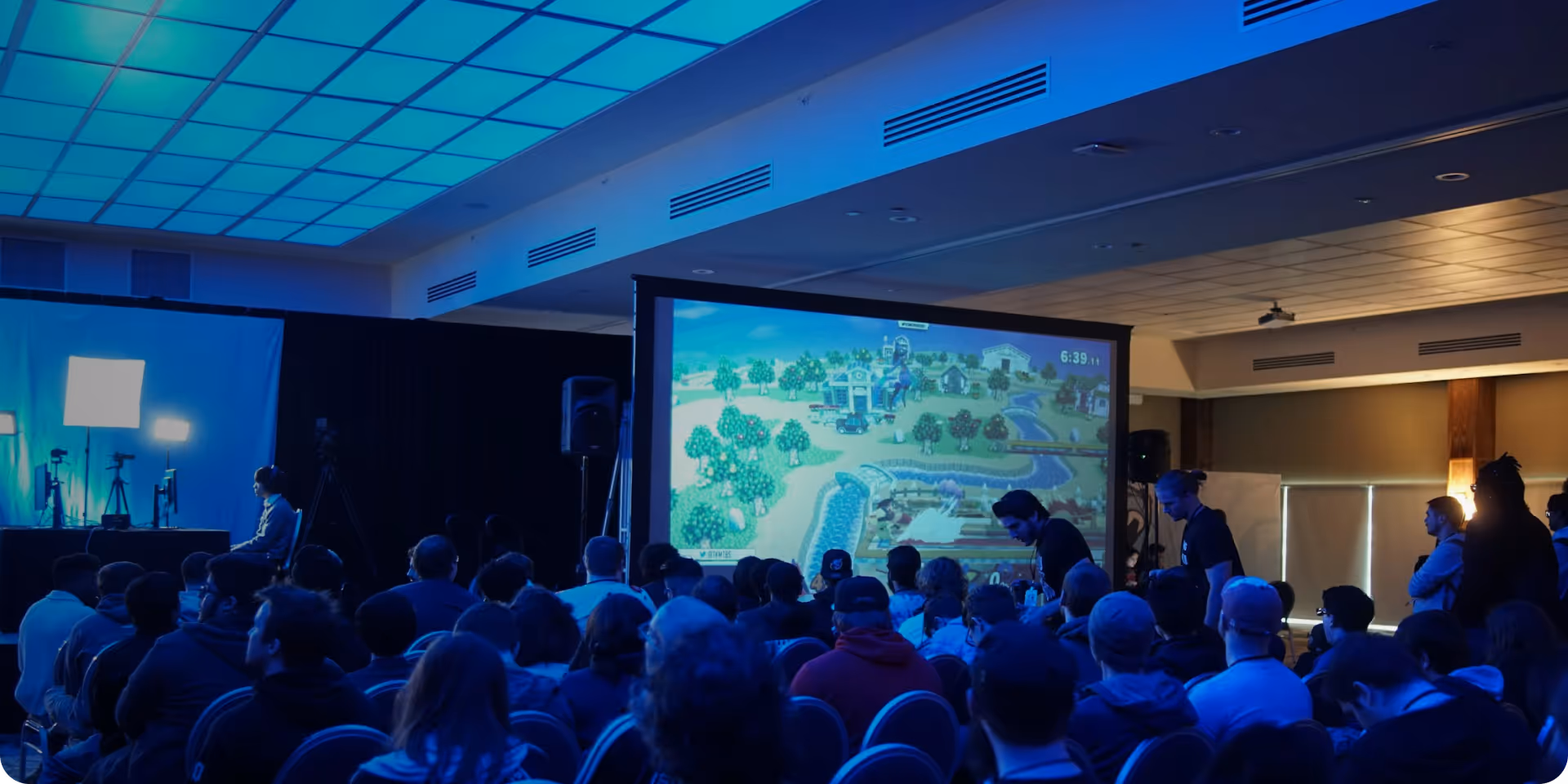 Audience watching a video game tournament on a large screen in a dimly lit room with gaming setup and lighting in the background.