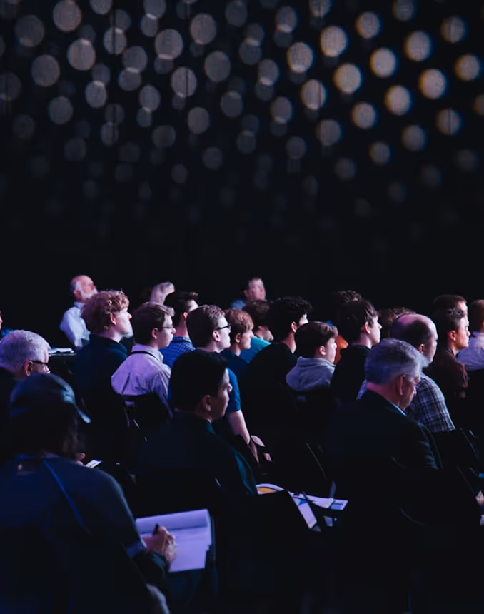 Audience attentively watching a presentation in a dark room with round light patterns on the ceiling.